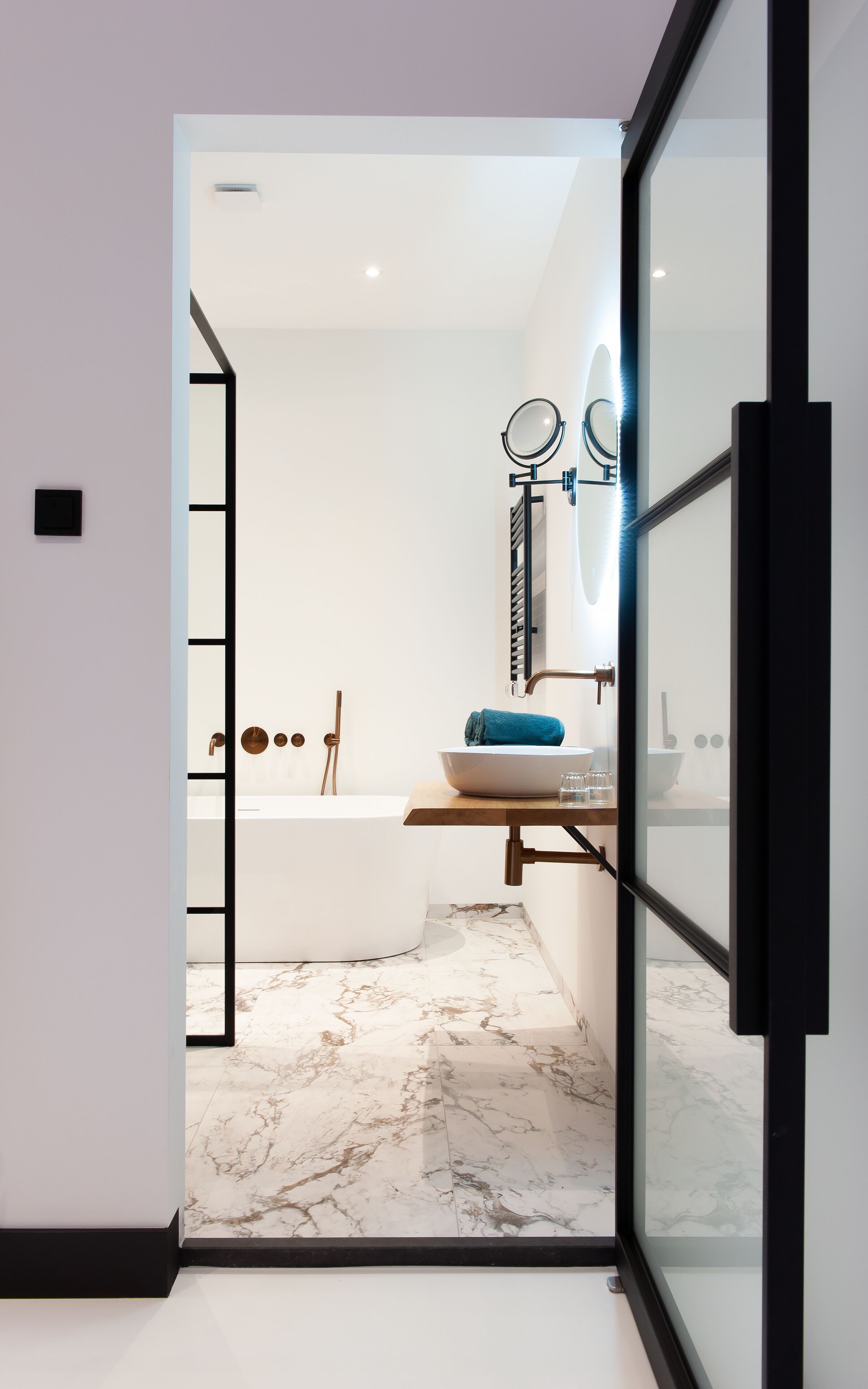 A modern bathroom with a marble floor, a bathtub, a sink with a blue towel, and a mirror with a light.