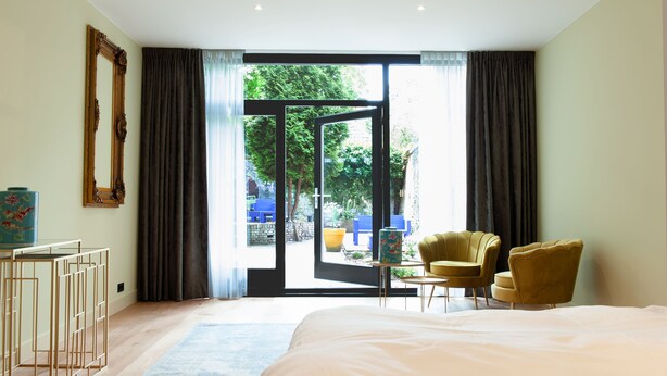 A bedroom with a large bed, a desk with a vase, two chairs, and a view of a garden through a glass door. - De Salon van Fagel (The Hague)