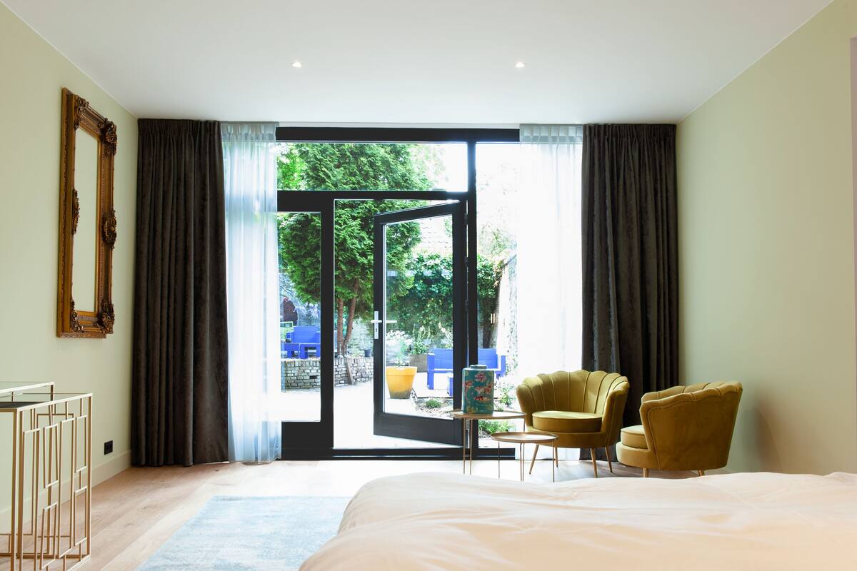 Ein Schlafzimmer mit einem großen Bett, einem Schreibtisch mit einer Vase, zwei Stühlen und Blick auf einen Garten durch eine Glastür.