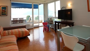 A living room with a sofa, a dining table, and a TV. There is a balcony with a view of the sea. - Lugano Blue Marine On Lake Shore (Lugano)
