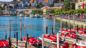 A waterfront area with red boats docked, a clear blue sky, and buildings in the background. - Lugano Blue Marine On Lake Shore (Lugano)