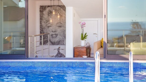 Una piscina interior moderna con cerramiento de vidrio, una pared decorativa con patrón de espiga y vistas al mar.