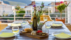 Una mesa de comedor con una botella de vino, copas, queso, aceitunas y flores en un balcón con vistas a la montaña.