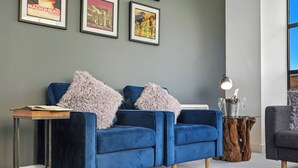 Una sala de estar moderna con un sofá azul, almohadas mullidas, una mesita de madera con una lámpara y cuadros en la pared.