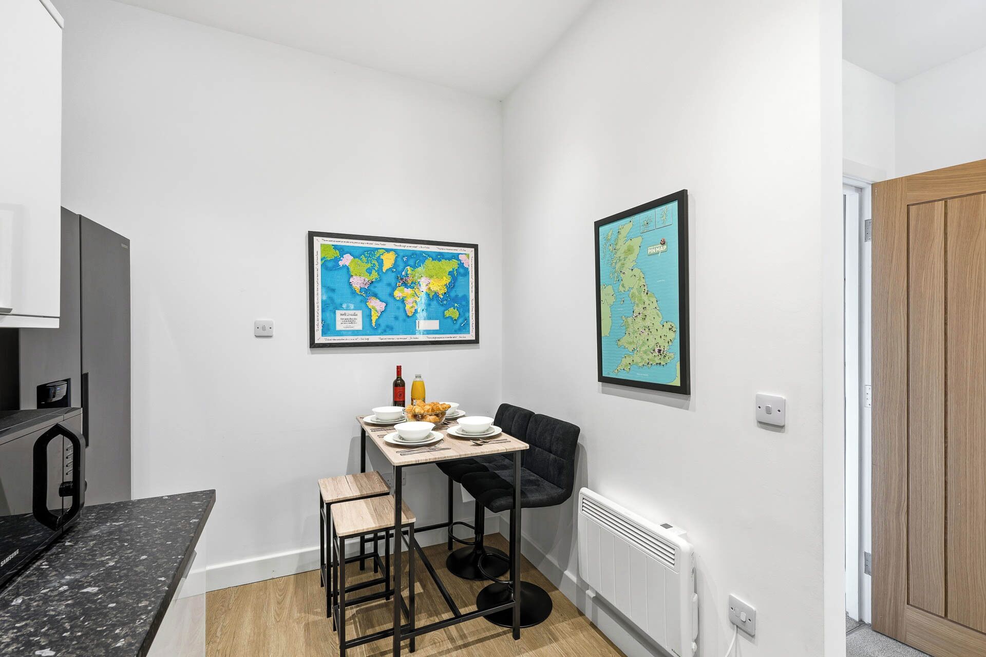 A small, modern kitchen with a dining area, featuring a map of the world and a map of the UK on the walls.