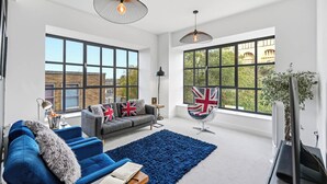 Amplia sala de estar con ventanas grandes, un sofá gris con banderas británicas, un sillón azul y una alfombra azul.