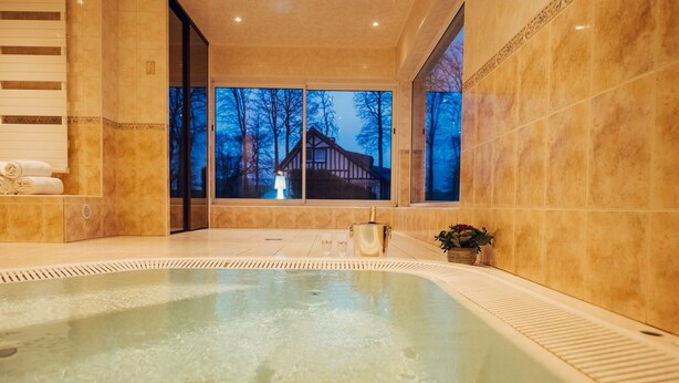 A spa area with a hot tub, a bottle of champagne, and a view of a house through large windows. - Clos Céleste - Campagne d'Etretat (Ecrainville)
