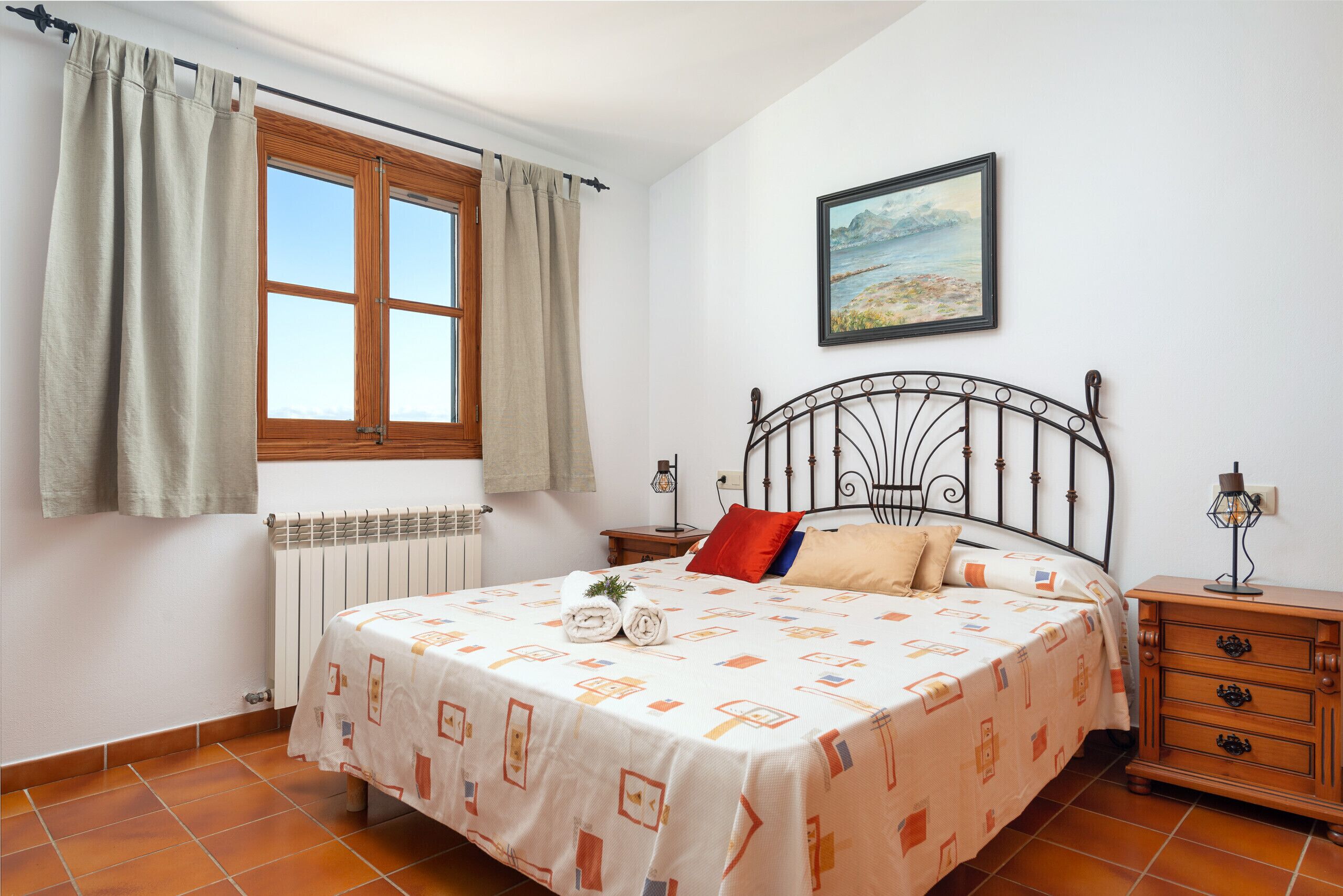 Un dormitori amb un llit, una finestra amb cortines, un radiador, una nitzeta amb una làmpada i un quadre a la paret.