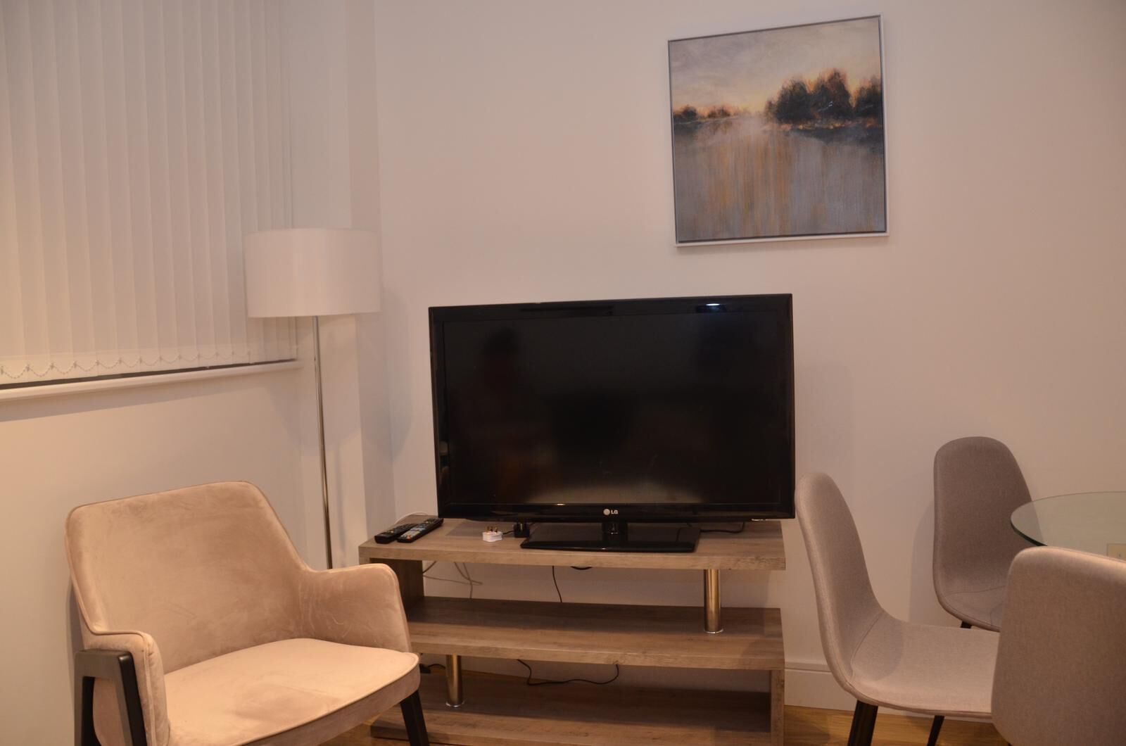 A living room with a flat-screen TV on a wooden stand, two beige armchairs, a glass coffee table, and a painting on the wall.