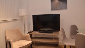 A living room with a flat-screen TV on a wooden stand, two beige armchairs, a glass coffee table, and a painting on the wall. - LARPOTE LODGE - LUTON (LUTON)
