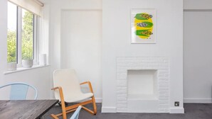 A room with a white brick fireplace, a framed abstract artwork, a wooden chair, a table, and a window with a view of greenery.