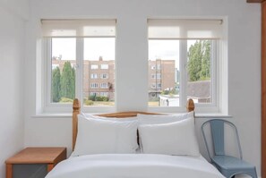 Ein Schlafzimmer mit einem Bett, zwei Fenstern, einem Sessel und einem kleinen Tisch.