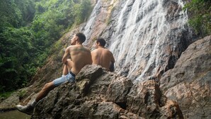 Deux personnes sont assises sur des rochers, près d’une cascade, dans une zone boisée.