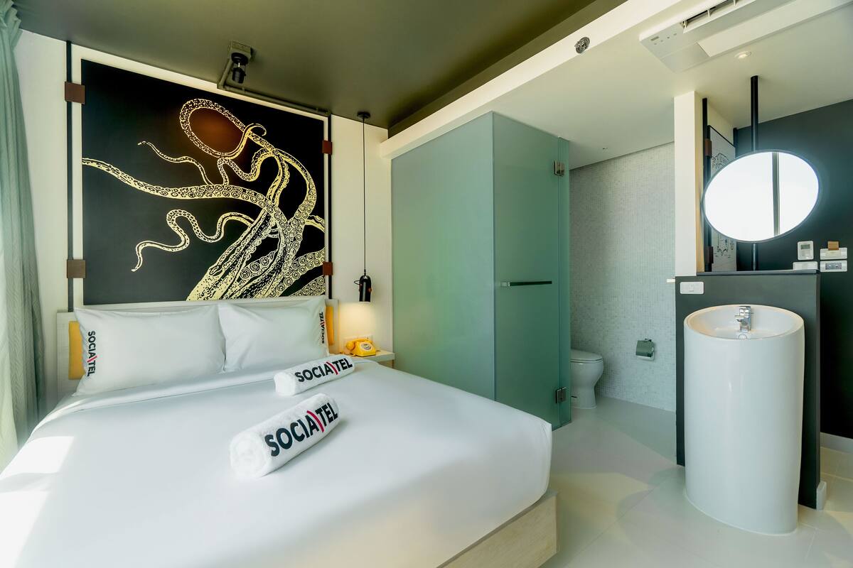 Une chambre d’hôtel comprenant un lit, une grande fresque murale, une salle de bain avec un lavabo et un miroir, ainsi que des toilettes.