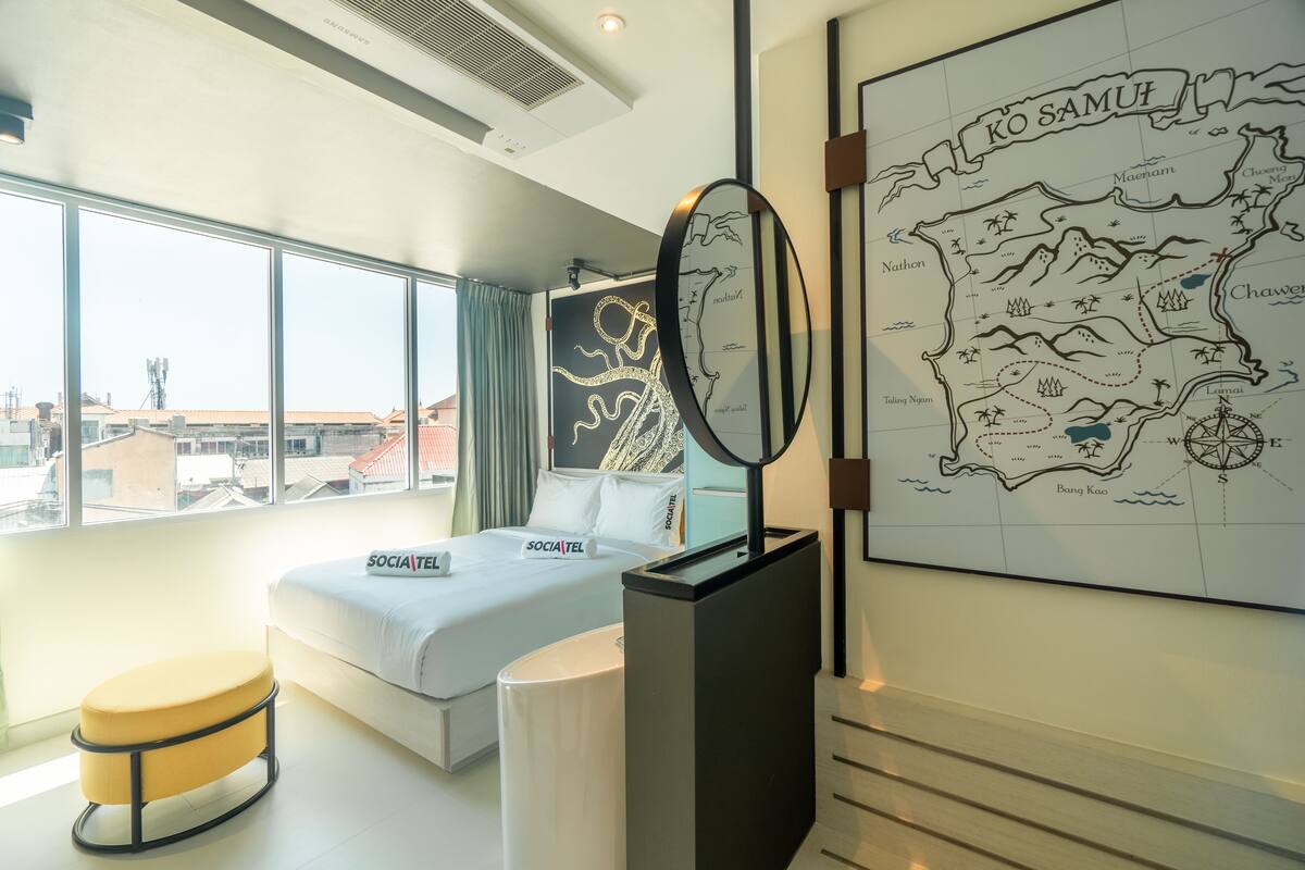 Une chambre d’hôtel moderne avec une grande fenêtre, un lit doté d’un oreiller « SOCIA TEL », une carte accrochée au mur et une petite table ronde.