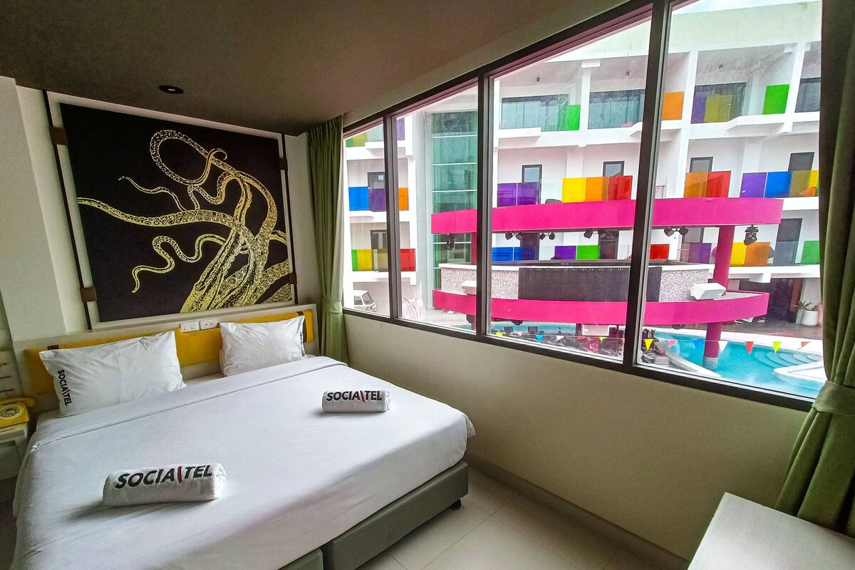 Une chambre d’hôtel avec un lit, une fenêtre décorée de motifs colorés et une vue sur un immeuble doté de balcons.