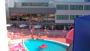 Un espace piscine doté de chaises longues et de parasols, entouré d’un bâtiment avec de grandes fenêtres et un balcon.