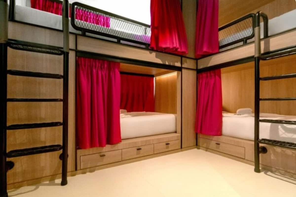 Une chambre avec des lits superposés, chacun doté d’une structure en bois et d  ’un matelas. Des rideaux sont accrochés aux fenêtres, et une échelle est fixée sur le côté des lits.