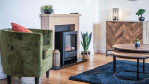 Een gezellige woonkamer met een groene fauteuil, een open haard, een houten kast en een ronde salontafel.