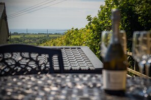 Une table avec une bouteille de vin et un verre, placée sur fond d’un paysage rural avec des champs et une vue lointaine sur la mer.