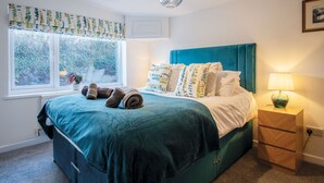 Een slaapkamer met een blauwgroene sprei, meerdere kussens, een bedlampje en een raam met gordijnen.