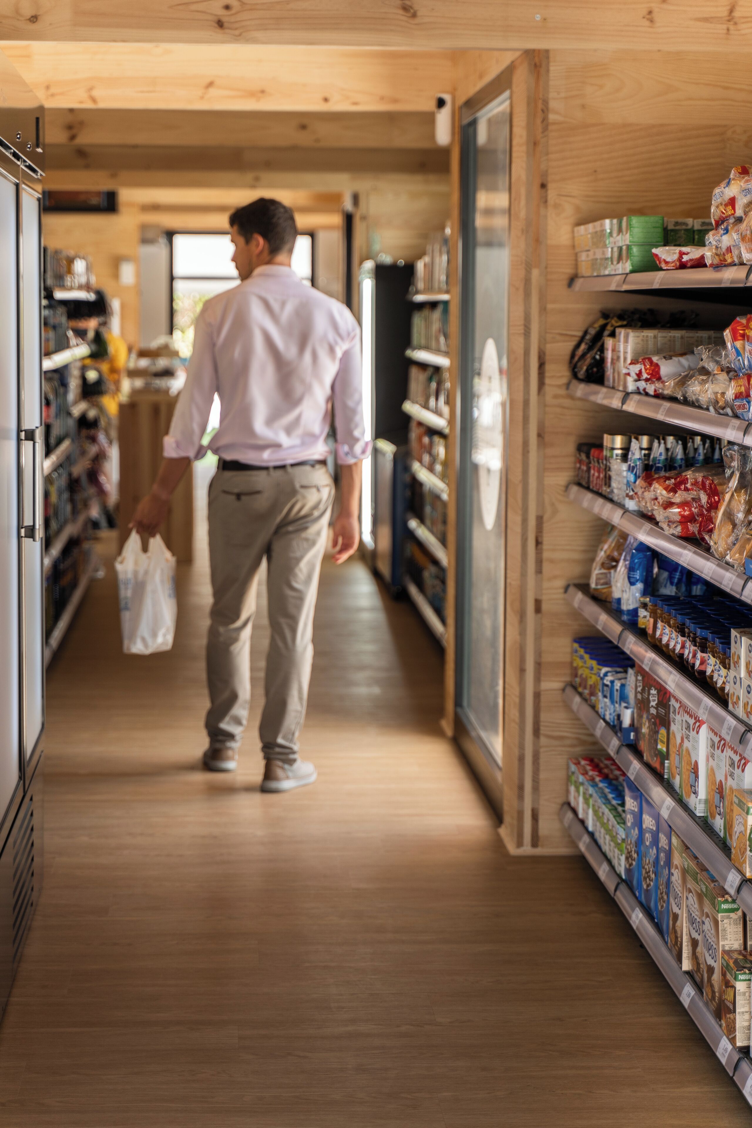 A man walking down a wooden-floored corridor in a store, shelves stocked with various products.