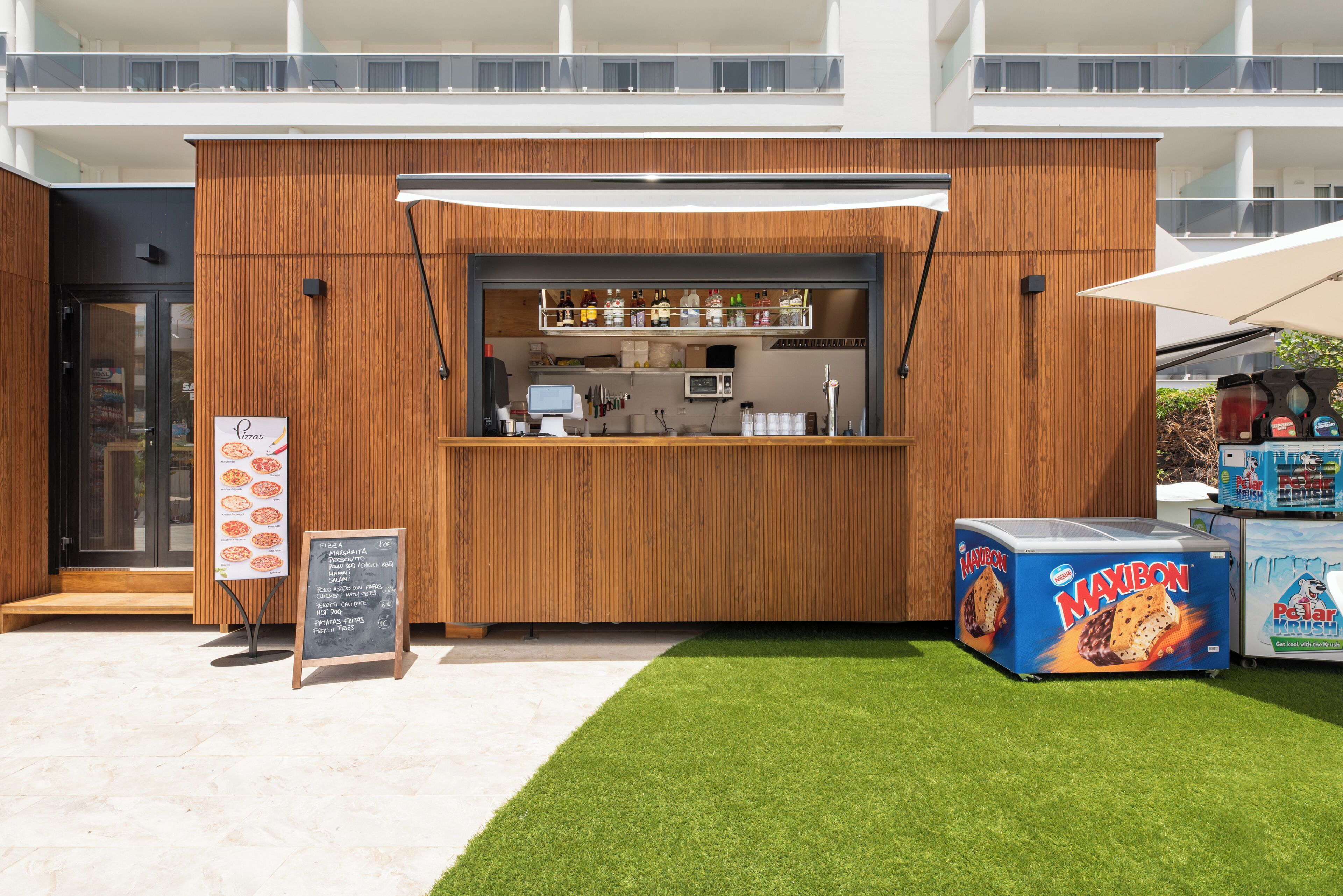 A modern outdoor food kiosk with a wooden facade, a menu board, and a Maxibon ice cream cooler.