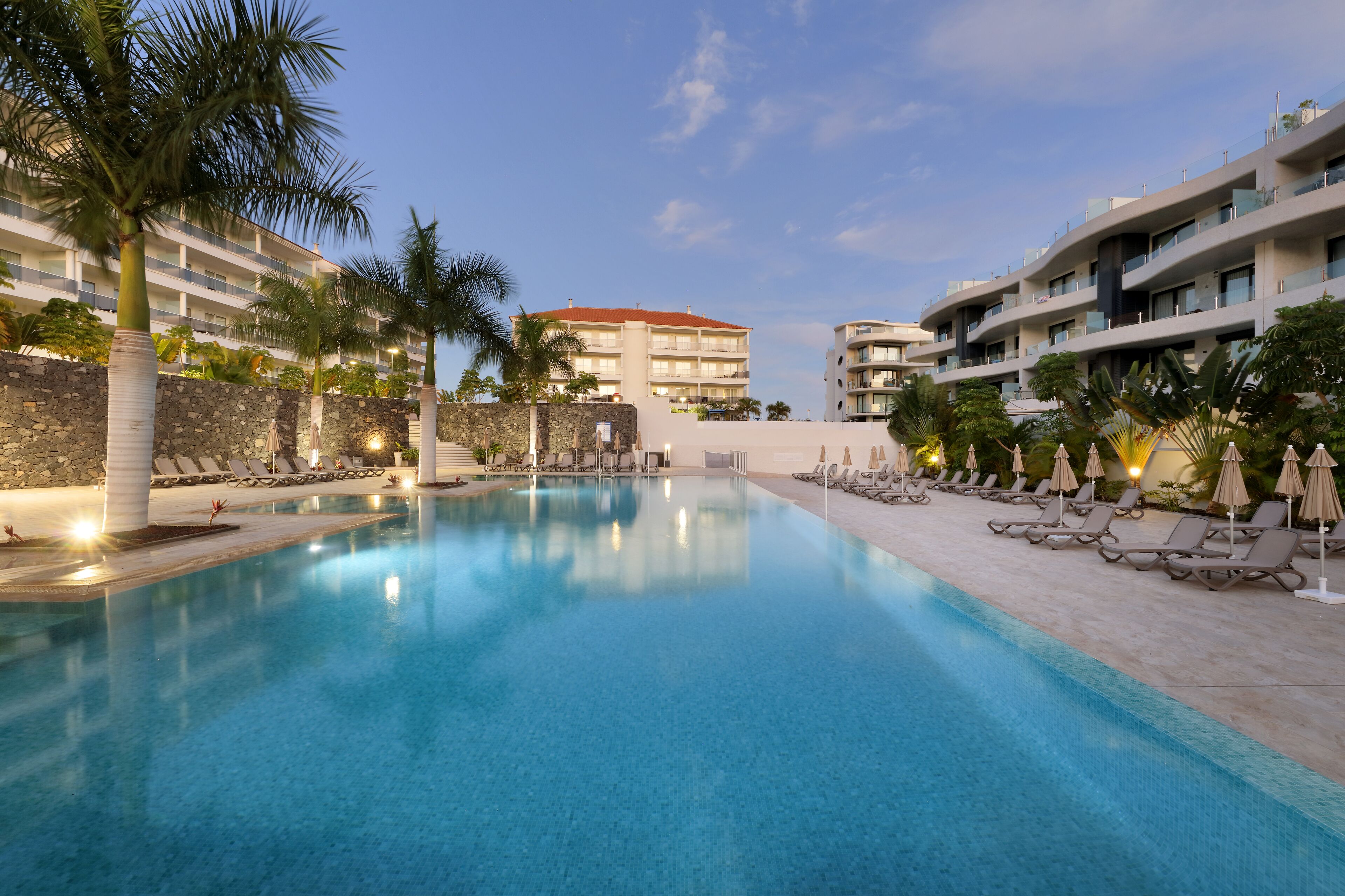 Un espace piscine d’hôtel avec de nombreux chaises longues et parasols, entouré de palmiers et d’un mur en pierre.