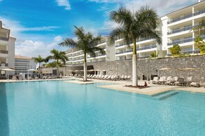 A swimming pool with lounge chairs and palm trees, surrounded by a multi-story building with balconies.