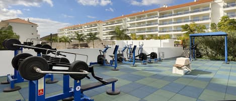 A gym with various exercise equipment, including weight machines and free weights, set against a backdrop of a multi-story building with balconies and palm trees.