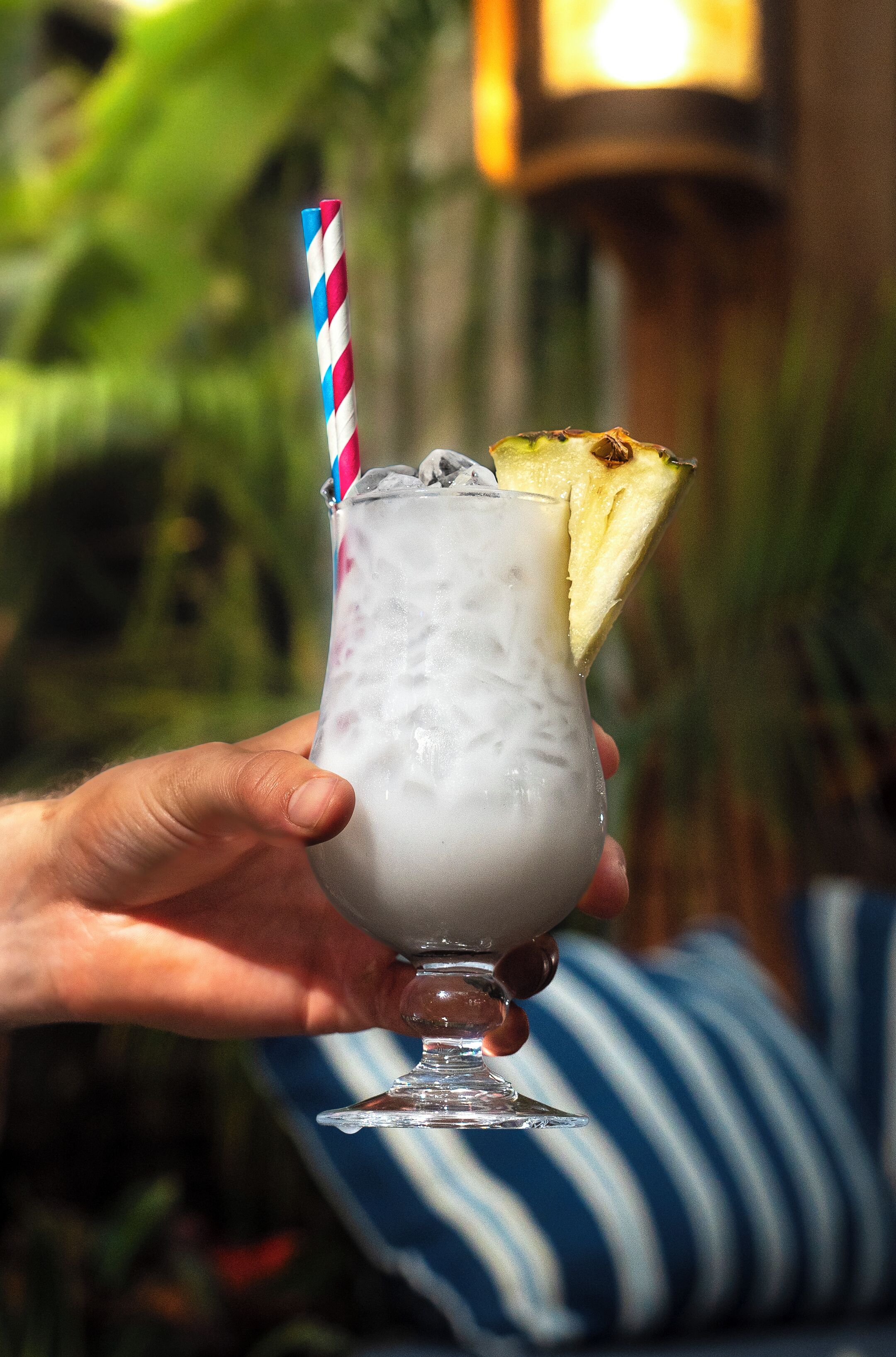 A hand holding a glass with a striped straw, a slice of pineapple, and ice cubes.