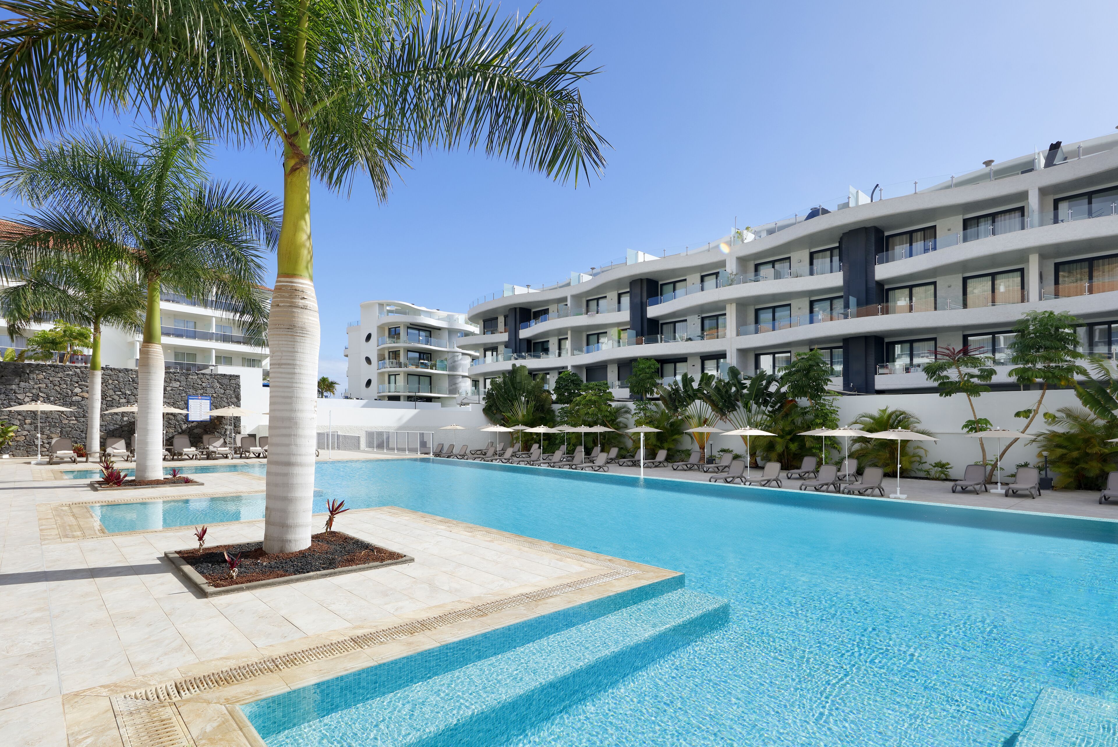 A swimming pool with lounge chairs and umbrellas, surrounded by palm trees and a modern building with balconies.