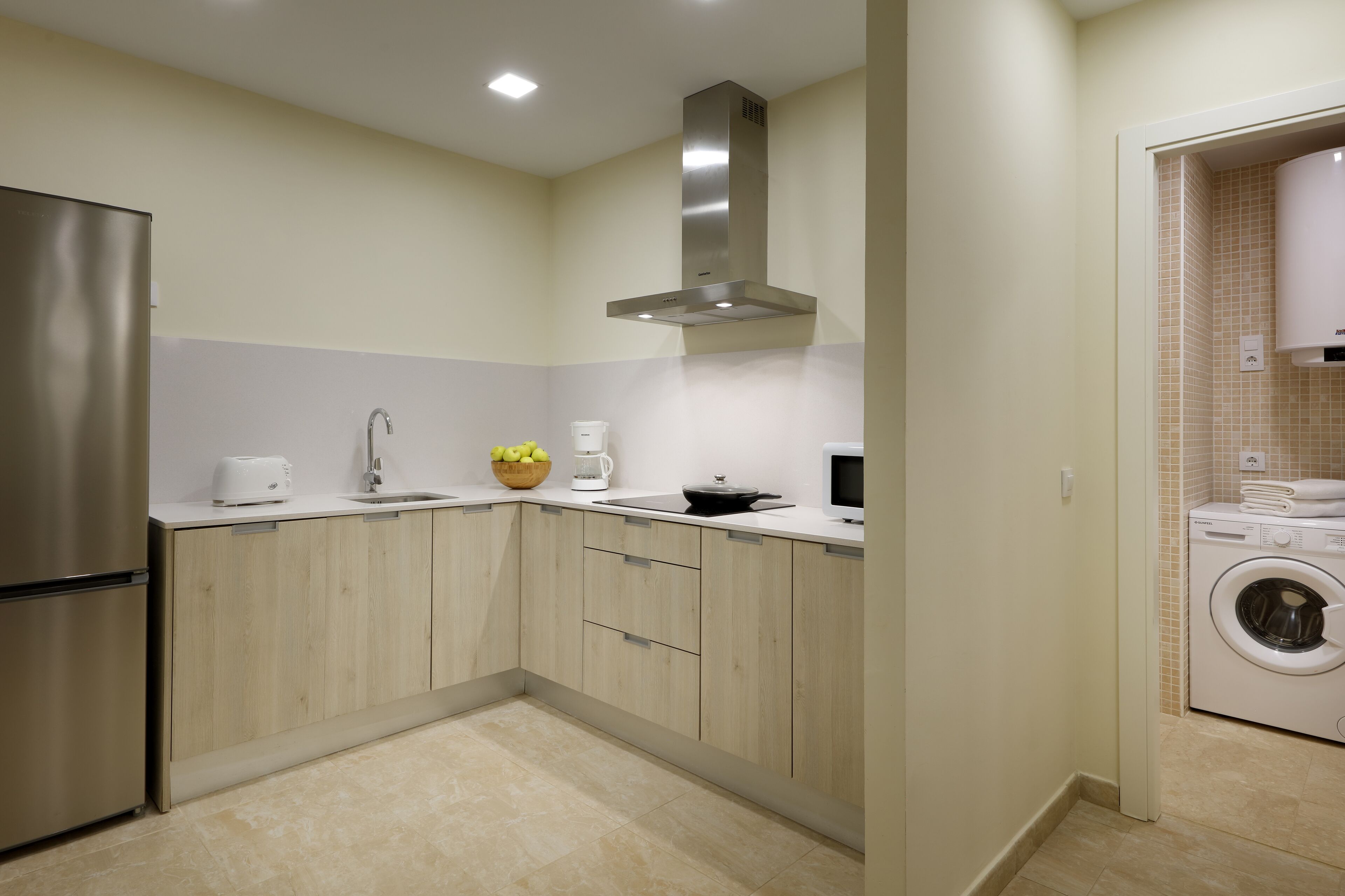 A modern kitchen with a stainless steel refrigerator, a microwave, a sink, and a stove. There is a washing machine in the adjacent room.