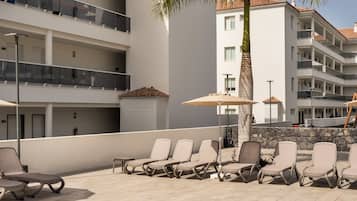Zona de piscina con sillas de descanso y una palmera, rodeada por un edificio de varios pisos con balcones.