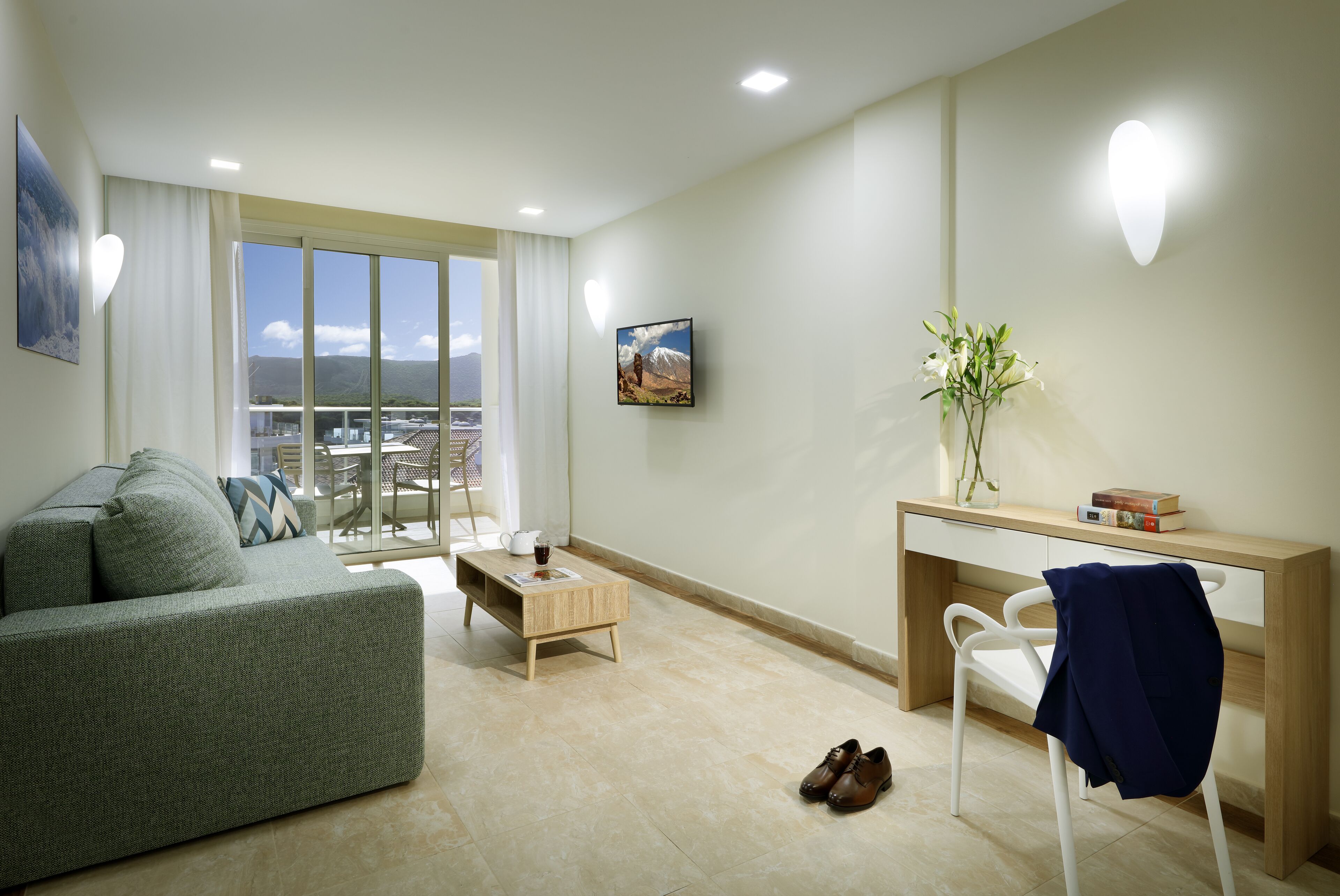 A modern living room with a sofa, coffee table, and a desk with a chair. There is a view of a cityscape through large windows.