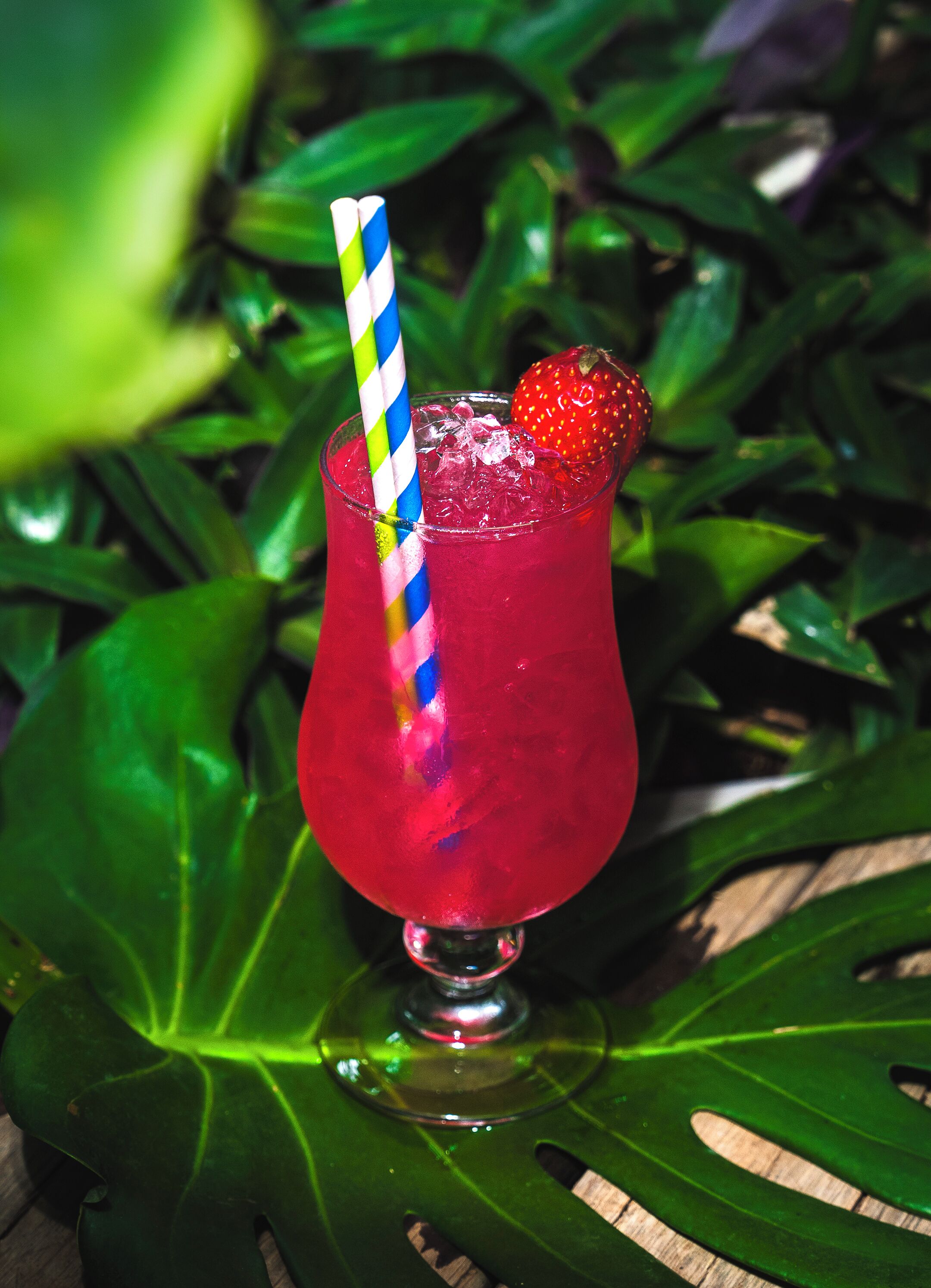 A glass of red beverage with a striped straw and a strawberry on the rim, placed on a green leaf.