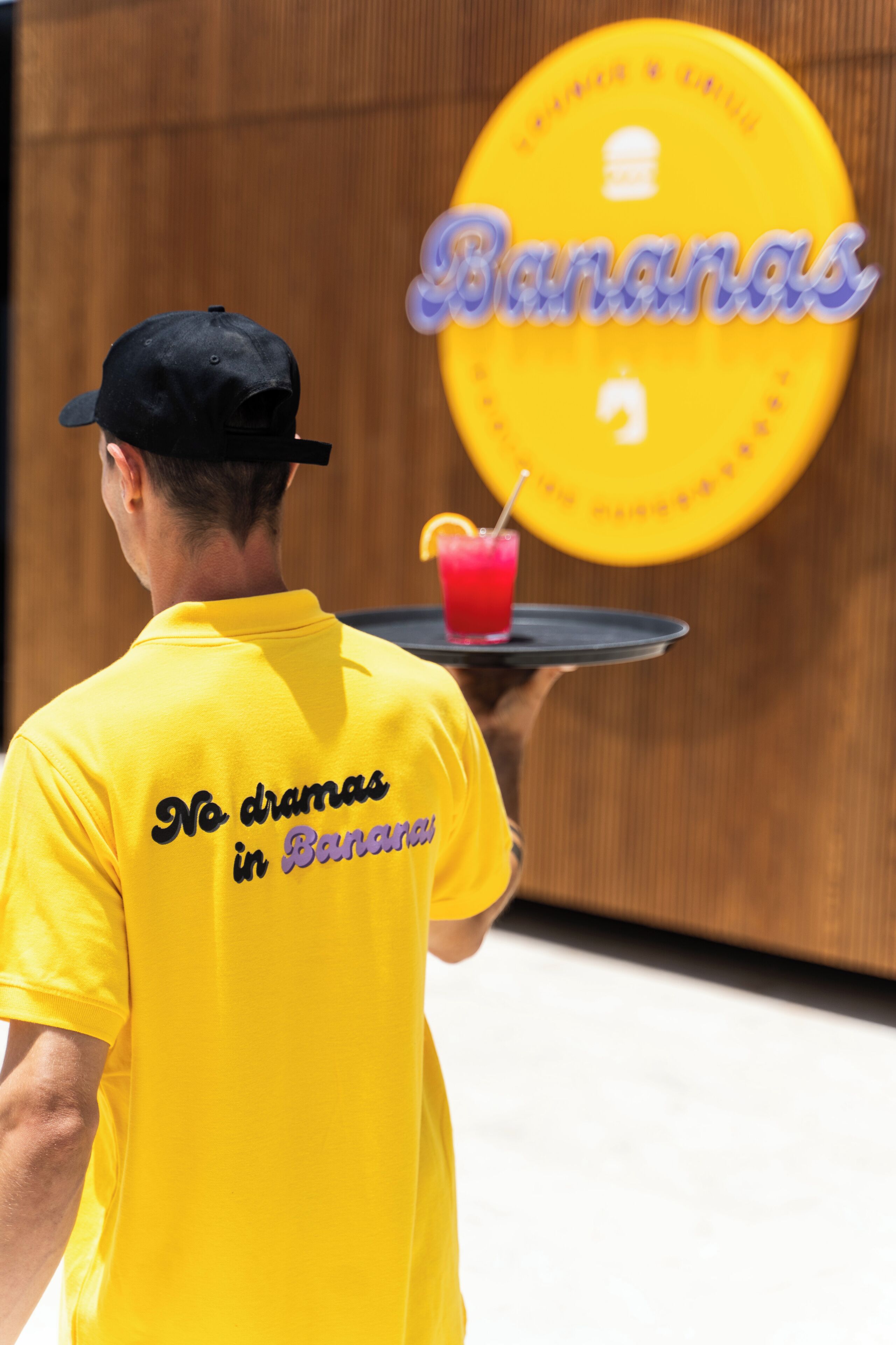 A person in a yellow shirt with "No dramas in Banana" text, holding a tray with a drink and a banana.