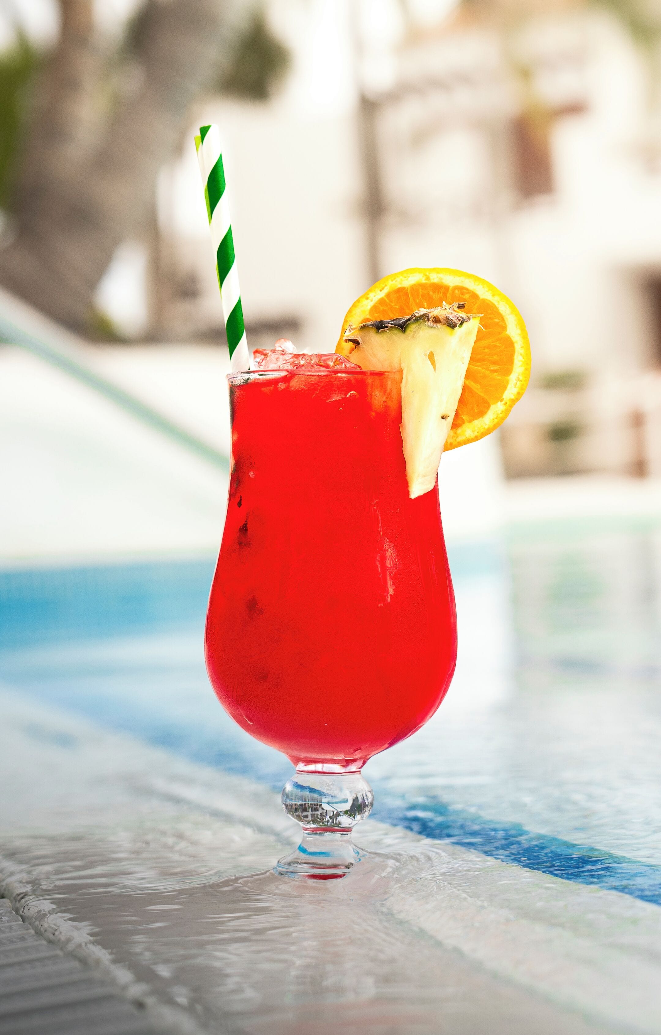 A red cocktail with a green and white striped straw and an orange slice garnish, served in a glass with a stem, placed on a reflective surface near a pool.