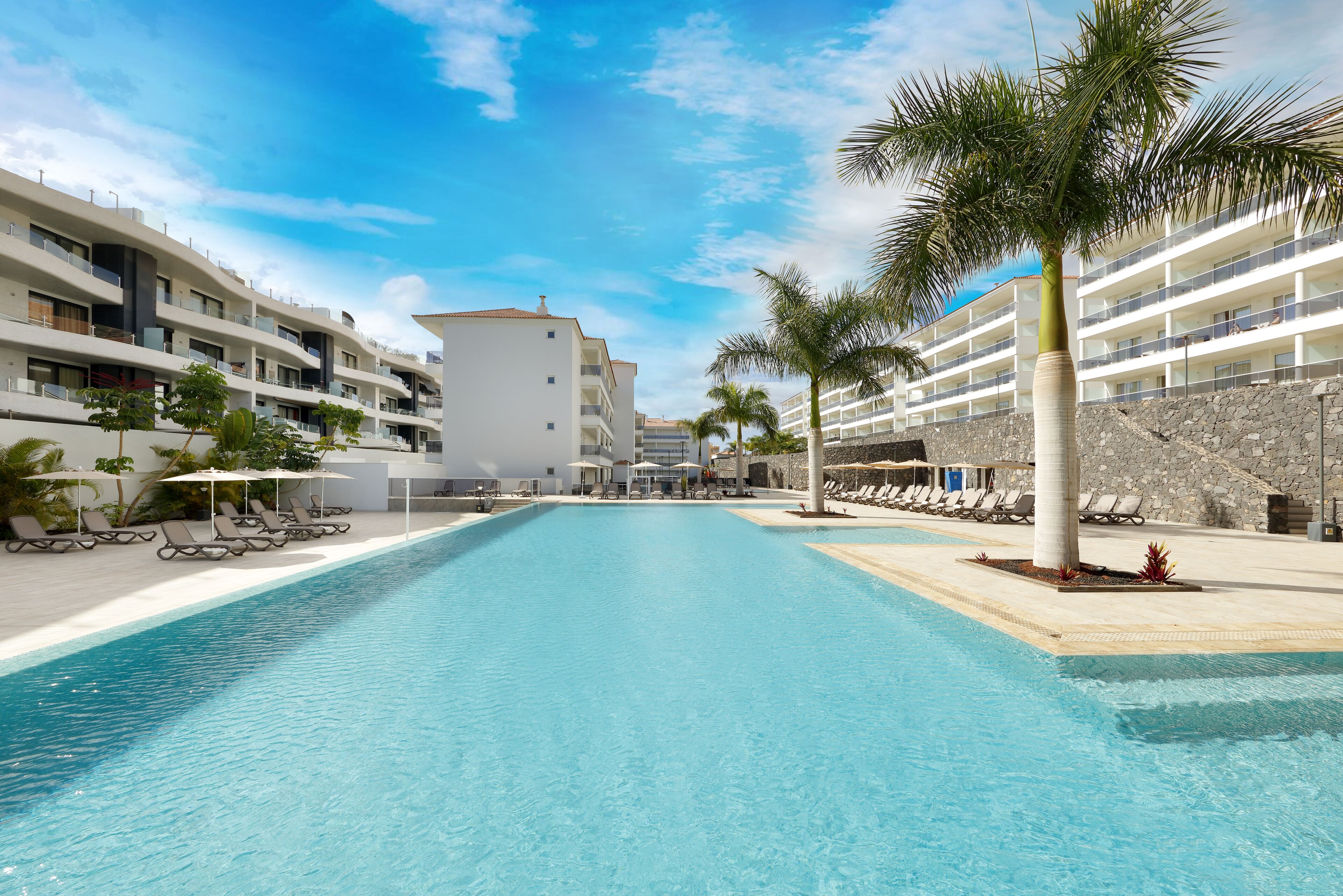 A swimming pool with lounge chairs and umbrellas, surrounded by a hotel building with balconies and palm trees.