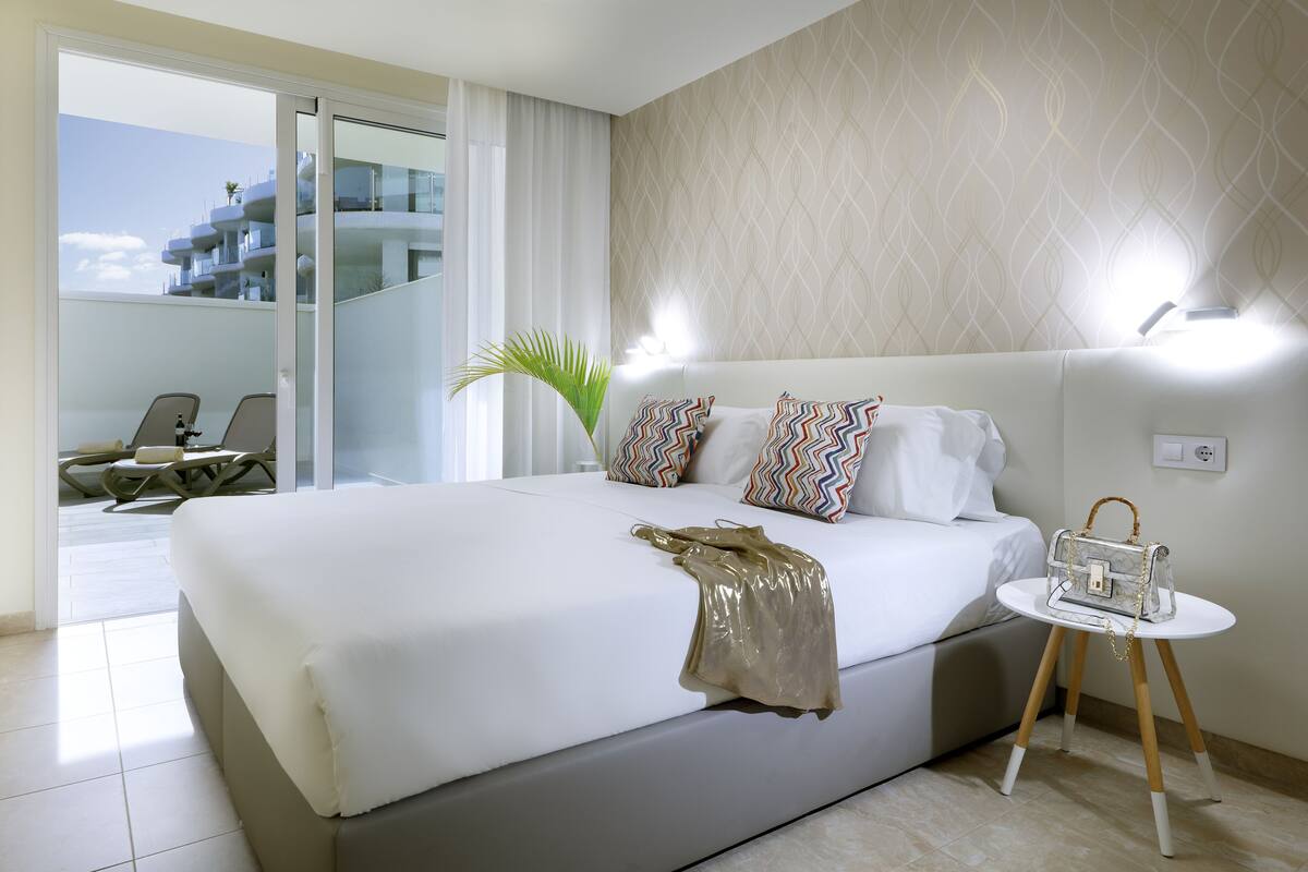 Een moderne hotelkamer met een groot bed, een klein tafeltje met een handtas, en uitzicht op een gebouw door een glazen deur.