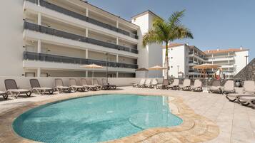 A swimming pool with lounge chairs and umbrellas, surrounded by a multi-story building with balconies.