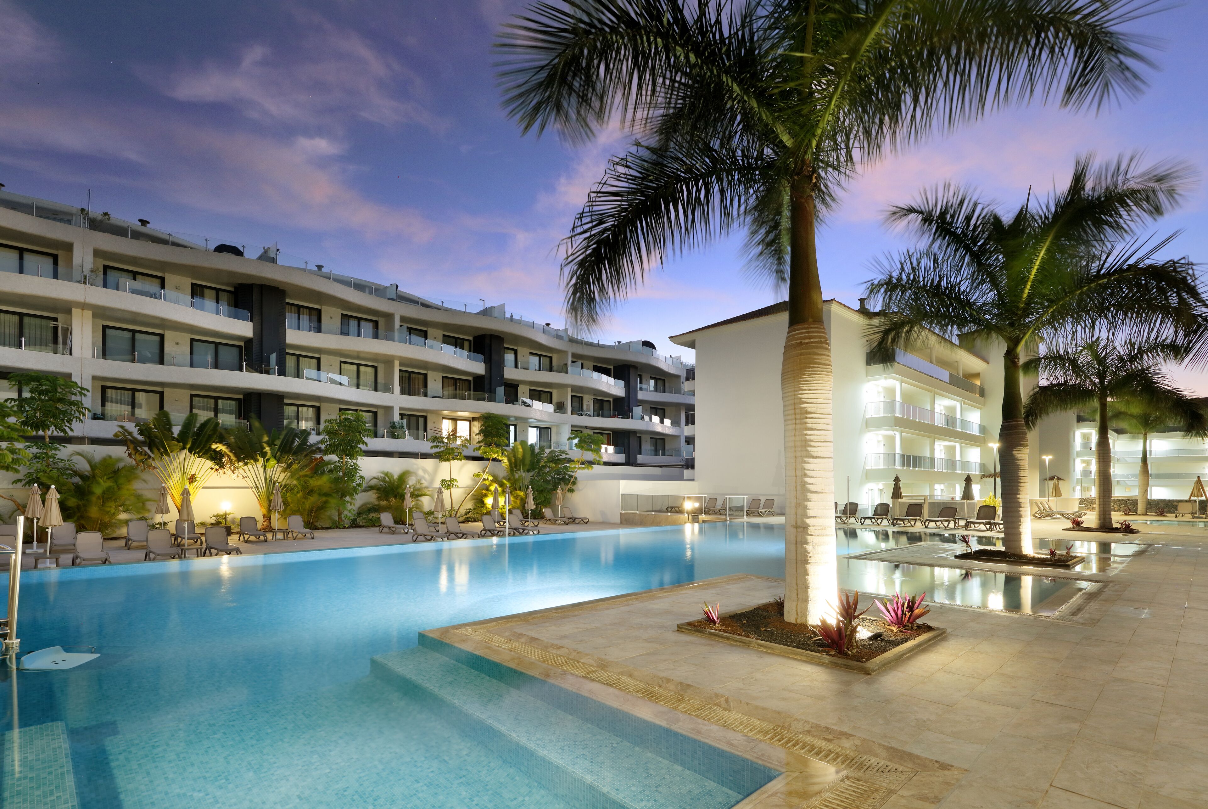Un hotel con una gran piscina, palmeras y sillones de descanso.