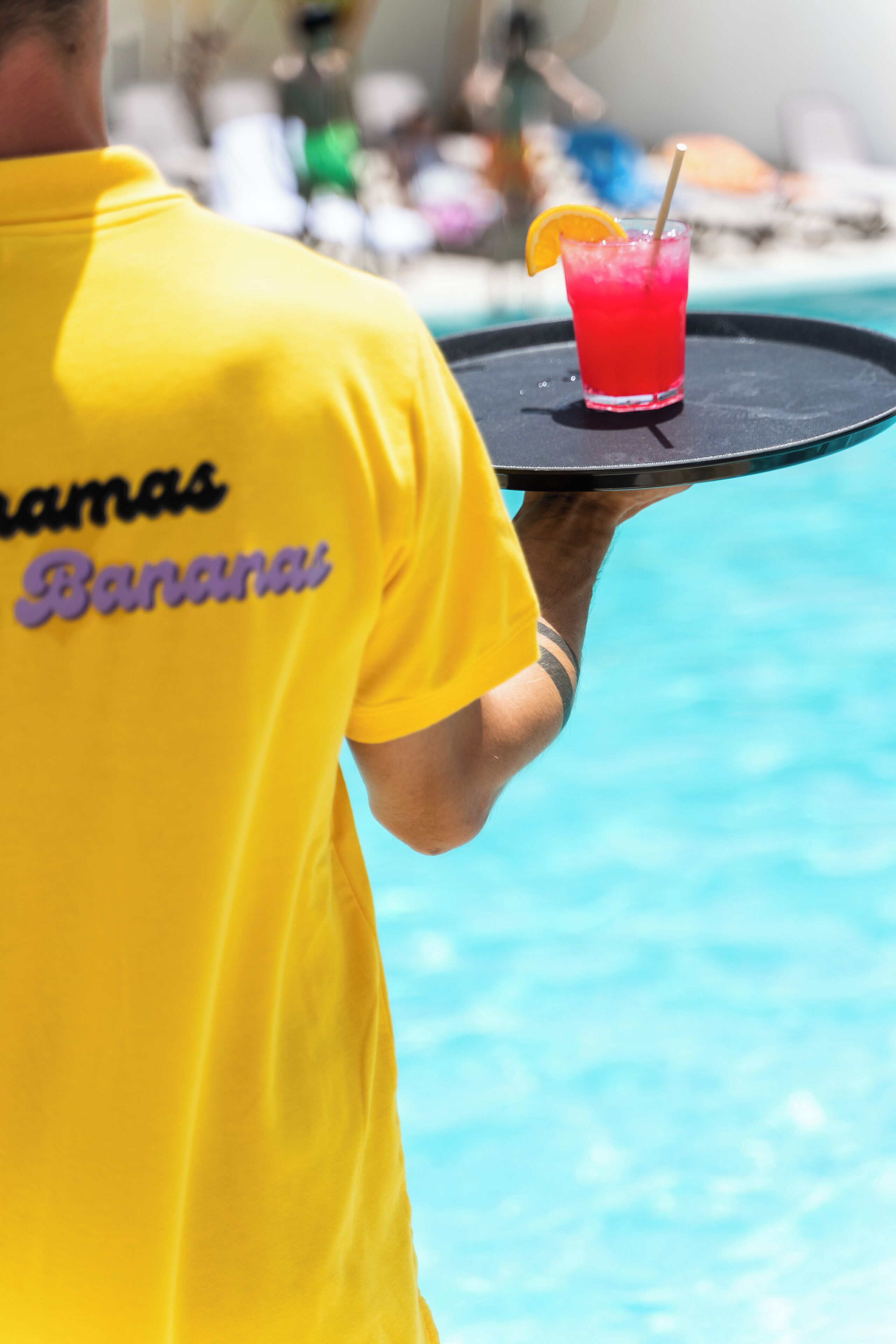 A person in a yellow shirt holding a tray with a red drink and an orange slice.