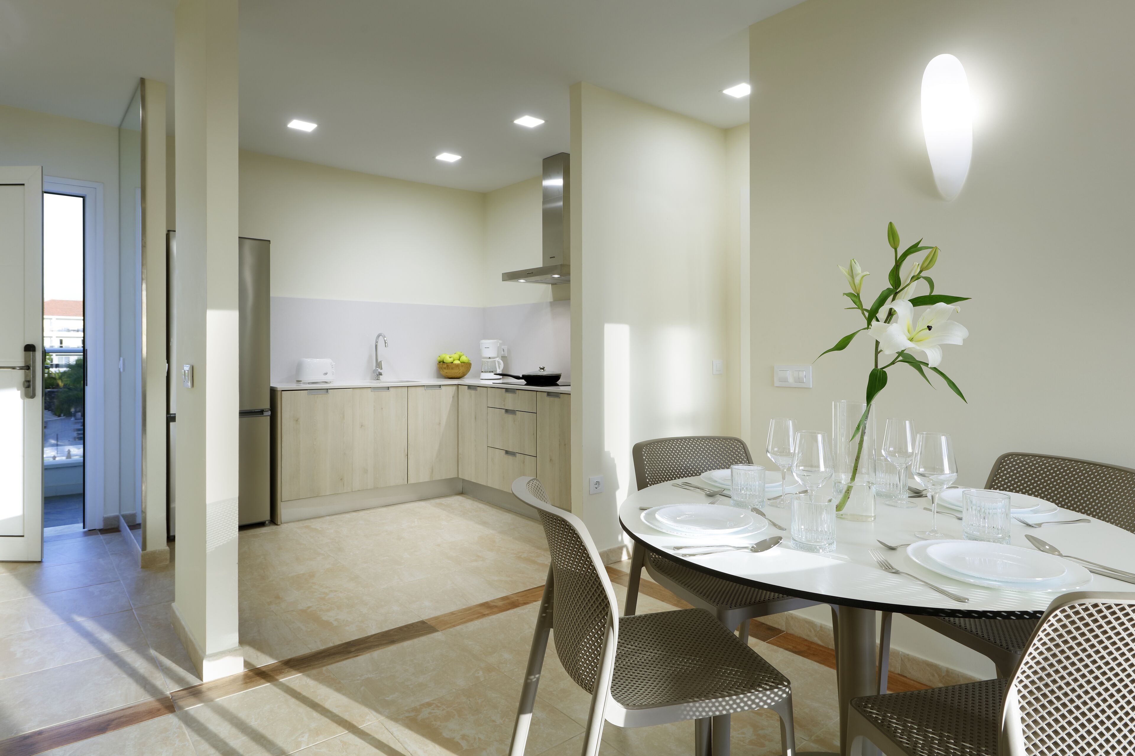 A modern dining area with a round table set for six, surrounded by six chairs. The room has a neutral color palette with beige walls and a tiled floor. There is a kitchen area visible in the background with wooden cabinets and a stainless steel range hood.
