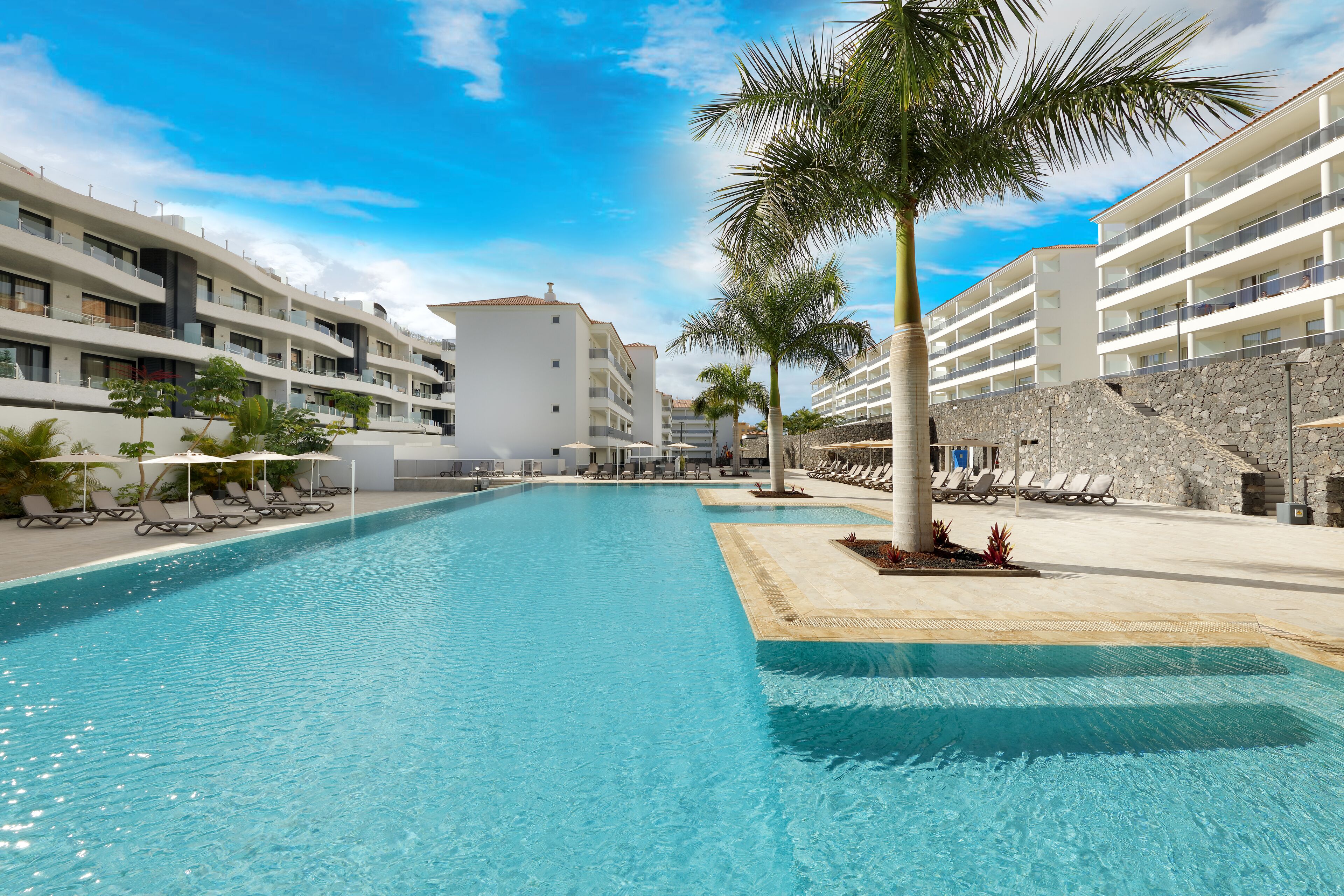 A swimming pool with lounge chairs and palm trees, surrounded by multi-story buildings with balconies.