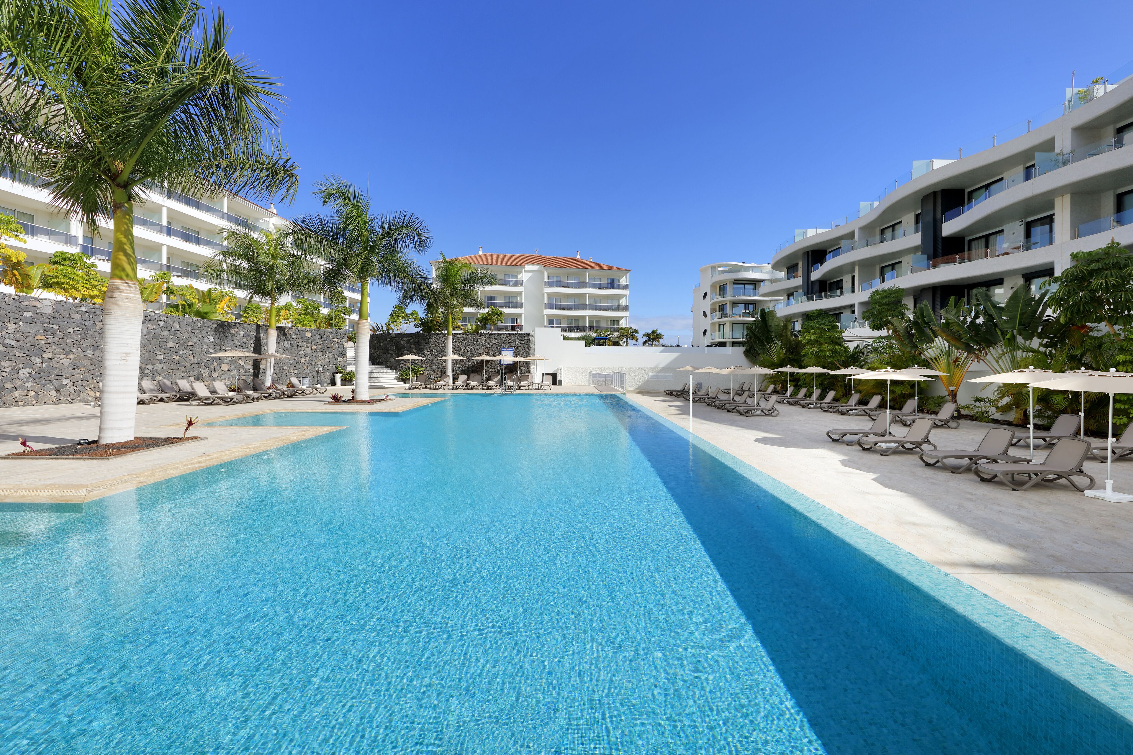 A swimming pool with lounge chairs and palm trees, surrounded by modern buildings with balconies.