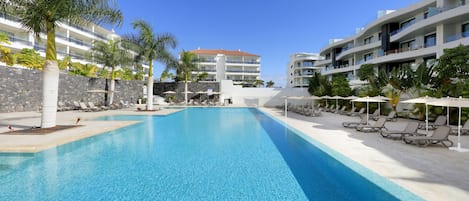 Une piscine entourée de chaises longues et de palmiers, le tout bordé de bâtiments modernes dotés de balcons.