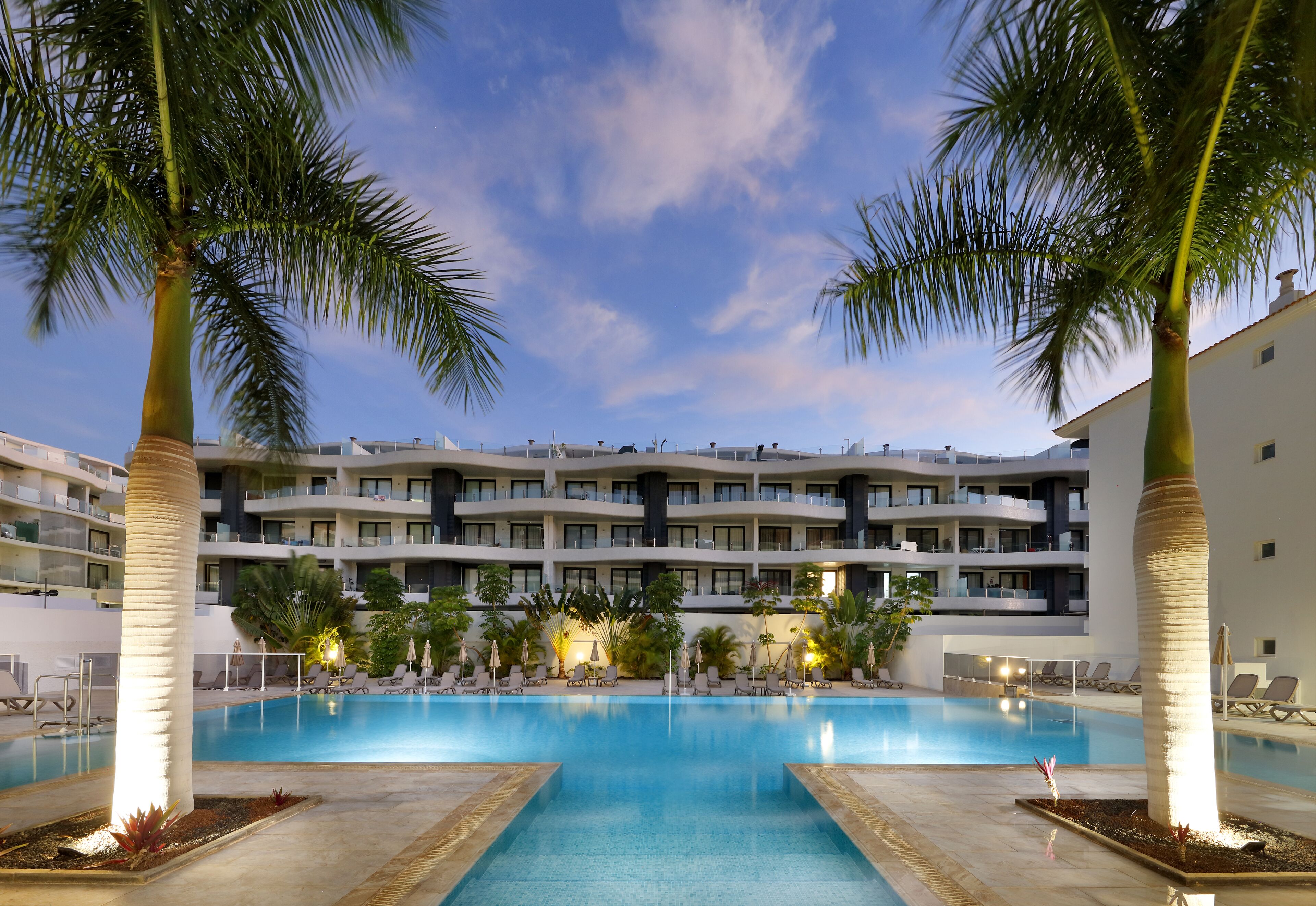 A swimming pool surrounded by palm trees and lounge chairs, with a multi-story hotel building in the background.