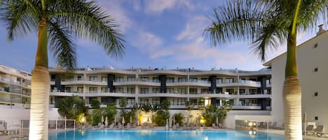A swimming pool surrounded by palm trees and lounge chairs, with a multi-story hotel building in the background.