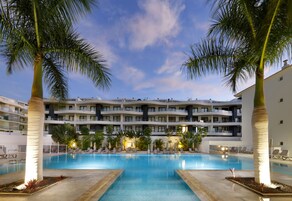 Piscina cercada por palmeiras e espreguiçadeiras, com um prédio de hotel de vários andares ao fundo.